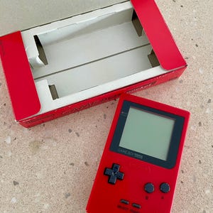 K&ouml;nnte beinhalten: Ein rotes Game Boy Pocket Handheld-Konsolen mit schwarzem Bildschirm und Kn&ouml;pfen, neben der roten Pappschachtel. Die Schachtel ist ge&ouml;ffnet und zeigt einen wei&szlig;en Innenraum. Auf der Konsole steht "GAME BOY pocket".