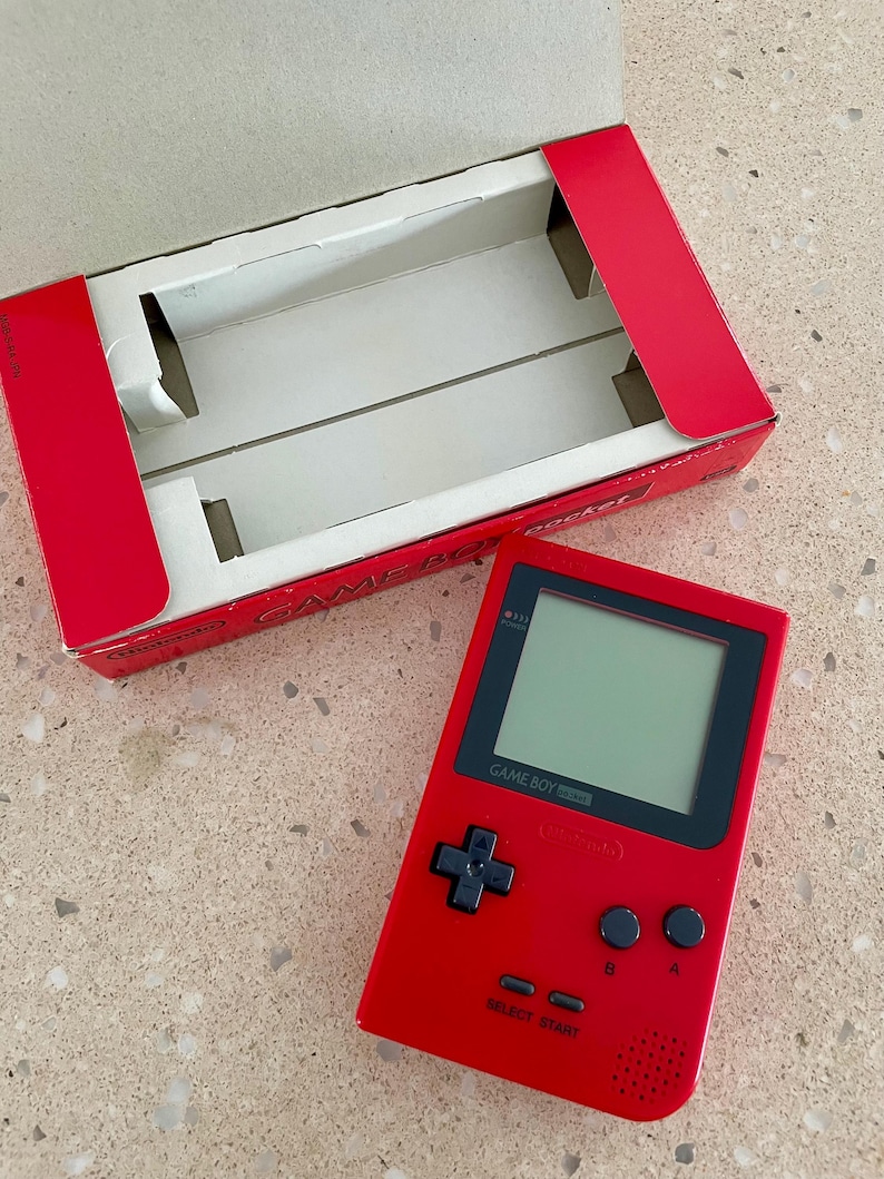 K&ouml;nnte beinhalten: Ein rotes Game Boy Pocket Handheld-Konsolen mit schwarzem Bildschirm und Kn&ouml;pfen, neben der roten Pappschachtel. Die Schachtel ist ge&ouml;ffnet und zeigt einen wei&szlig;en Innenraum. Auf der Konsole steht "GAME BOY pocket".