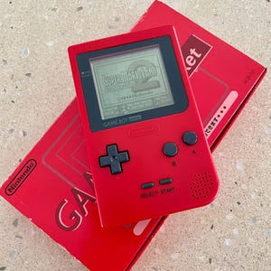 May include: A red Game Boy Pocket console with the game Super Mario Land displayed on the screen. The console is next to its red box, which has the Nintendo logo and the word "GAME" printed on it.