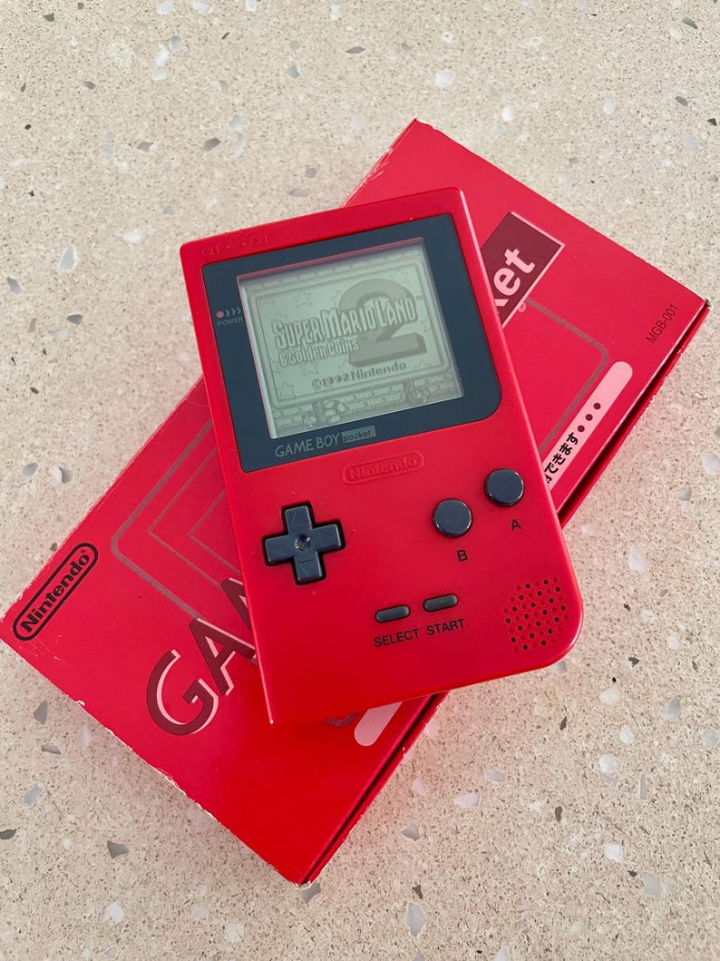 K&ouml;nnte beinhalten: Ein rotes Game Boy Pocket-Konsolen-Ger&auml;t mit dem Spiel Super Mario Land auf dem Bildschirm. Die Konsole befindet sich neben der roten Box mit dem Nintendo-Logo und dem Wort "GAME".