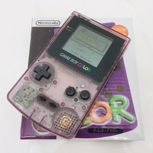 Puede incluir: Una consola de juegos portátil Game Boy Color de color púrpura transparente con una pantalla negra que muestra "Super Mario Land". La consola está encima de su caja original, que presenta el logotipo de Nintendo y las palabras "Game Boy Color".