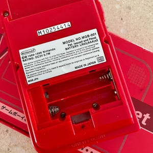K&ouml;nnte beinhalten: Ein rotes Nintendo Game Boy MGB-001, das Batteriefach und die R&uuml;ckseite zeigend. Das Ger&auml;t hat die Seriennummer M10254414 und enth&auml;lt Text, der darauf hinweist, dass es in Japan hergestellt wurde. Das Ger&auml;t ben&ouml;tigt LR03(AAA)x2 Batterien.