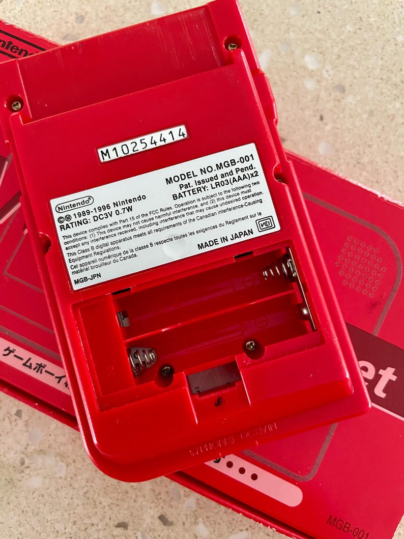 K&ouml;nnte beinhalten: Ein rotes Nintendo Game Boy MGB-001, das Batteriefach und die R&uuml;ckseite zeigend. Das Ger&auml;t hat die Seriennummer M10254414 und enth&auml;lt Text, der darauf hinweist, dass es in Japan hergestellt wurde. Das Ger&auml;t ben&ouml;tigt LR03(AAA)x2 Batterien.