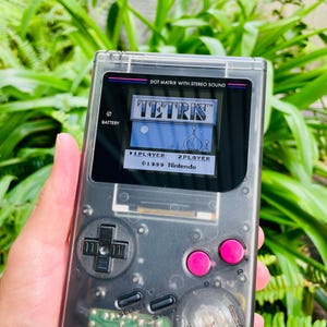May include: A transparent gray handheld gaming device displaying the game Tetris. The device has a directional pad, pink buttons, and the text "DOT MATRIX WITH STEREO SOUND". The screen shows the game's title and options for 1 or 2 players. The text "©1989 Nintendo" is also visible.