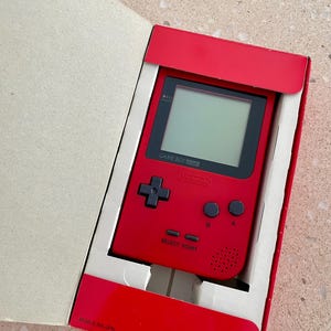 K&ouml;nnte beinhalten: Ein rotes Game Boy Pocket Handheld-Videospiel in der Originalverpackung. Die Konsole verf&uuml;gt &uuml;ber einen schwarzen Bildschirm, ein Steuerkreuz und Tasten. Die Verpackung ist rot und wei&szlig;.