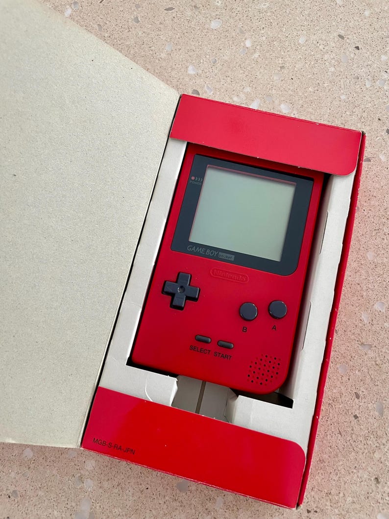 K&ouml;nnte beinhalten: Ein rotes Game Boy Pocket Handheld-Videospiel in der Originalverpackung. Die Konsole verf&uuml;gt &uuml;ber einen schwarzen Bildschirm, ein Steuerkreuz und Tasten. Die Verpackung ist rot und wei&szlig;.