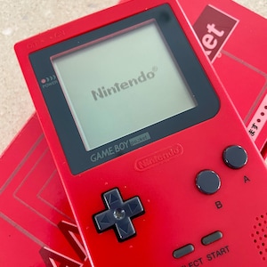 K&ouml;nnte beinhalten: Ein rotes Game Boy Pocket Handheld-Spielkonsolen mit einem schwarzen Bildschirm, auf dem das Nintendo-Logo angezeigt wird. Die Konsole hat schwarze Kn&ouml;pfe und ein Steuerkreuz. Eine rote Box mit dem Wort "GAME" ist teilweise sichtbar.