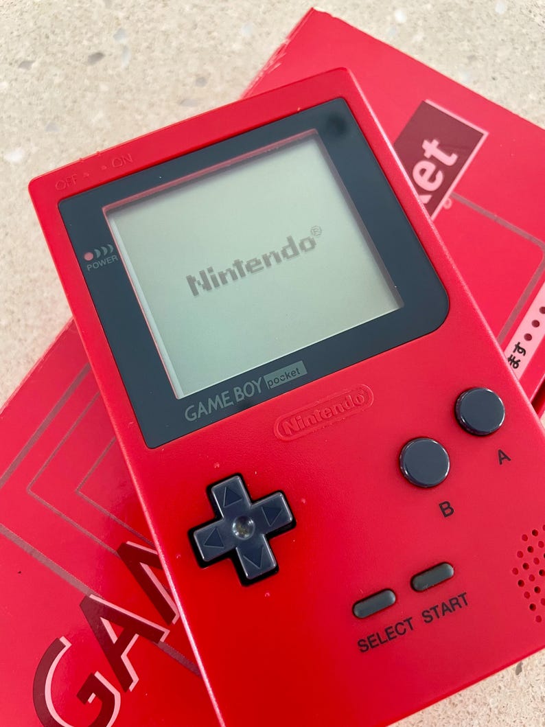 K&ouml;nnte beinhalten: Ein rotes Game Boy Pocket Handheld-Spielkonsolen mit einem schwarzen Bildschirm, auf dem das Nintendo-Logo angezeigt wird. Die Konsole hat schwarze Kn&ouml;pfe und ein Steuerkreuz. Eine rote Box mit dem Wort "GAME" ist teilweise sichtbar.