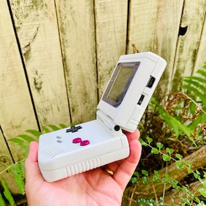 Puede incluir: Consola de videojuegos portátil blanca con pantalla abatible. La consola tiene un mando direccional, botones rosas y un altavoz. La pantalla es negra con un borde oscuro. La consola se sostiene en una mano.