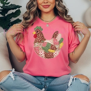 May include: A pink t-shirt featuring a patchwork chicken design. The chicken is made of various patterned fabrics, including floral, gingham, and heart-shaped patterns. The shirt is a solid pink color.