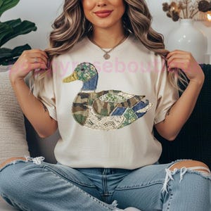 May include: Cream-colored t-shirt featuring a patchwork duck design. The duck has a green and blue head, a yellow beak, and a body composed of various fabric patterns, including camouflage and lace. The shirt is worn by a person.