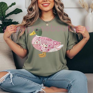 May include: An olive green t-shirt featuring a patchwork duck design. The duck has a green plaid head, a yellow beak, and pink and white patterned body with floral and heart motifs. The shirt is paired with blue jeans.