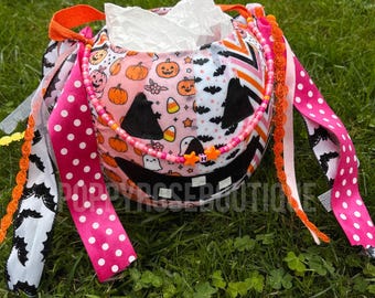May include: A fabric pumpkin bucket featuring a patchwork pattern of orange pumpkins, ghosts, bats, and candy corn. The bucket is decorated with long, hanging ribbons in pink, white, and black patterns, and a beaded handle with orange star charms.