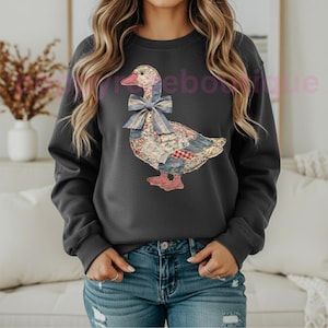 May include: Dark gray sweatshirt featuring a patchwork goose design with a blue bow. The goose is made of various fabric patterns, including floral and plaid. The sweatshirt is a casual, comfortable style.
