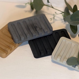 May include: A set of four rectangular soap dishes in tan, dark grey, black, and light beige. Each dish features a series of parallel raised ridges designed for drainage, with a smooth, solid construction and a matte finish throughout.
