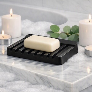 May include: A rectangular black soap dish with a slatted design holds a solid off-white bar of soap. The dish is surrounded by two large white pillar candles, two small silver tea light candles, and a sprig of green eucalyptus leaves.