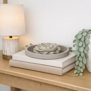 May include: A table lamp with a white fabric shade and a textured white and wood base, a gray circular tray containing a sculpted gray flower, and a white vase with trailing green leaves, all arranged on a stack of two books on a wooden table.