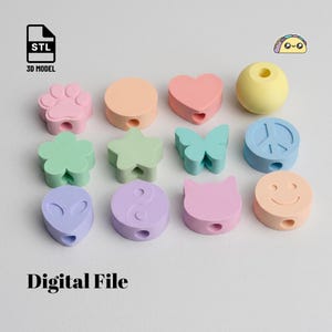 Op de afbeelding: Een verzameling van twaalf pastelkleurige kralen in diverse vormen, waaronder een pootafdruk, cirkel, hart, bol, bloem, ster, vlinder, vredesteken, alienhoofd, yin-yang, kattenkop en een smiley, elk voorzien van een gat om te rijgen.