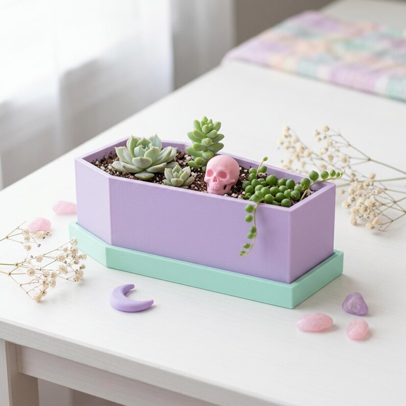 A light purple coffin-shaped planter box with a mint green tray sits on a white table filled with succulents, a small pink skull, and scattered crystals.