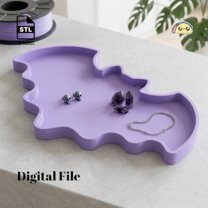 May include: A light purple tray shaped like a bat, designed as a 3D model digital file. The tray holds two small iridescent skull-shaped earrings, three dark purple crystal points, and a thin silver-toned chain necklace on its flat surface.