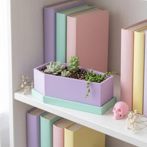 A light purple coffin-shaped planter box with a mint green tray is displayed on a white shelf among colorful books and a small pink skull figure.