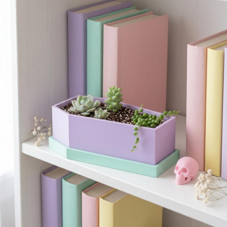 A light purple coffin-shaped planter box with a mint green tray is displayed on a white shelf among colorful books and a small pink skull figure.