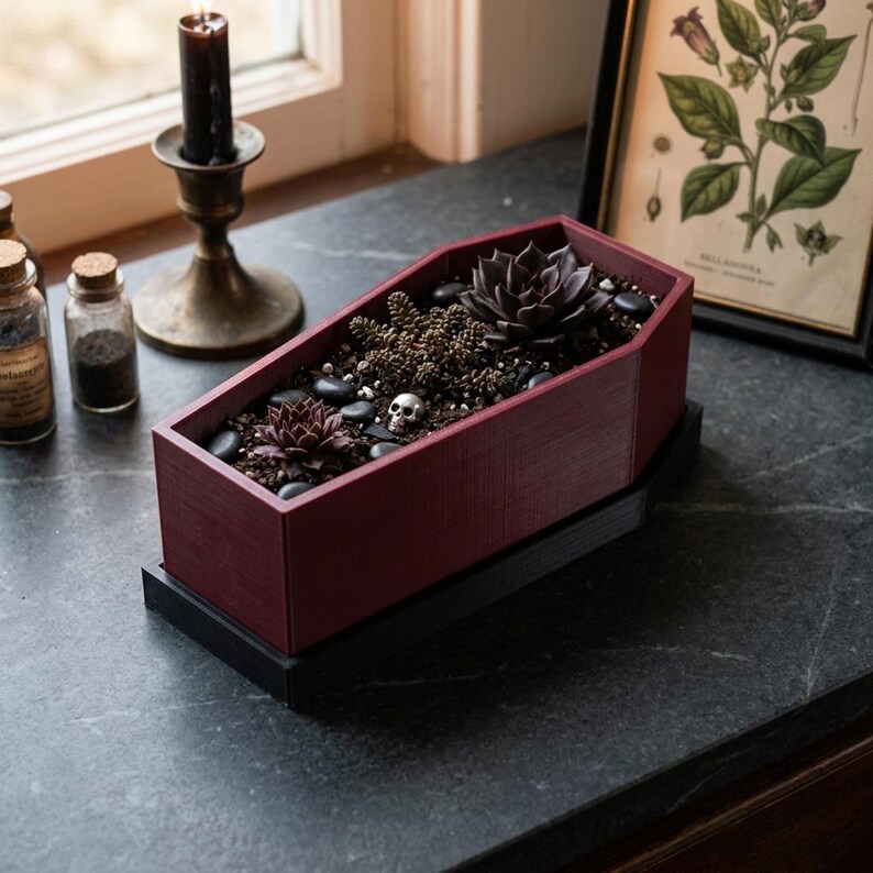 A dark red coffin-shaped planter with a black base, filled with dark succulents and small pebbles, sits on a dark surface next to a lit candle in a holder and a framed botanical print.