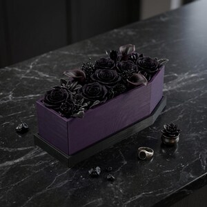 A purple coffin-shaped planter with a black base, filled with black roses, succulents, and small black pebbles, sits on a dark marble surface next to a ring and small black decorative items.