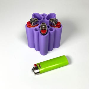 Puede incluir: Un soporte de plástico impreso en 3D de color lavanda con cinco ranuras cilíndricas, que contiene tres encendedores con detalles en naranja y rojo. Un encendedor Bic verde lima yace horizontalmente frente al soporte.