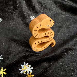 Puede incluir: Un juguete de serpiente de madera con un cuerpo curvado y una cara sonriente. La serpiente está hecha de madera de color claro y tiene detalles grabados. El juguete está sobre un fondo negro con purpurina en forma de copo de nieve.