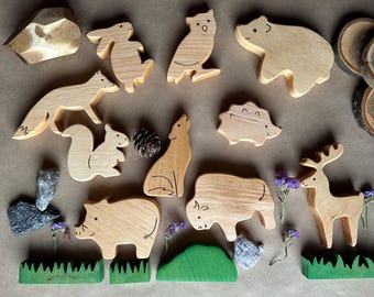 May include: Wooden animal figurines including a fox, squirrel, owl, bear, and deer. The set also includes a rock, a pine cone, and green grass blocks. The figurines are made of light-colored wood and are arranged on a brown surface.