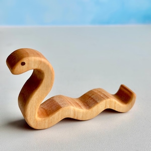 May include: A wooden toy snake, light brown in colour, with a simple, carved design. The snake is curled in an S-shape, with its head raised slightly.