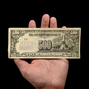May include: A vintage Japanese government 500 peso banknote with a green and brown color scheme. The note features a building with a tall chimney and the text "THE JAPANESE GOVERNMENT" and "FIVE HUNDRED PESOS".