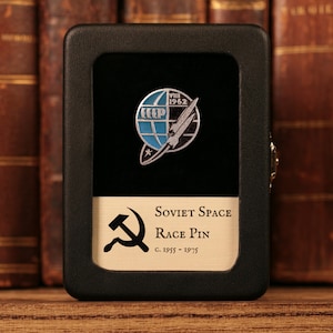 May include: A vintage Soviet Space Race pin in a black display case. The pin features a stylized rocket and globe design with blue accents. The case has a gold-coloured plaque with text and the hammer and sickle symbol.