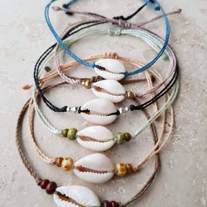 Könnte beinhalten: Ein Set aus sechs bunten Armbändern aus Schnur mit Kaurischnecken-Anhängern. Die Armbänder sind übereinander gelegt, wobei das oberste Armband blau, das zweite beige, das dritte hellgrün, das vierte schwarz, das fünfte grün und das unterste braun ist.