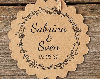 Könnte beinhalten: Ein braunes, gewelltes Papieretikett mit einem schwarzen floralen Kranzdesign. Der Text "Sabrina & Sven 05.08.21" ist innerhalb des Kranzes gedruckt.