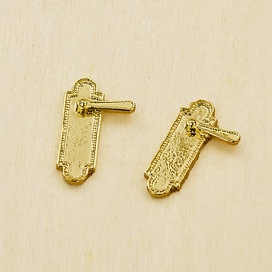 May include: Two small, gold-colored door handles with a rectangular shape and ornate detailing. Each handle has a small lever. The handles are set against a light-colored wooden surface.
