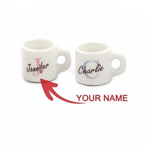 Pode incluir: Duas canecas de cerâmica branca com alças. A caneca à esquerda tem o nome "Jennifer" com um "J" rosa. A caneca à direita tem o nome "Charlie" com um "C" azul.