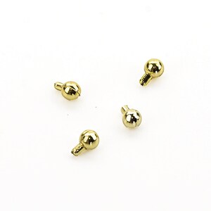 Könnte beinhalten: Vier goldfarbene Metallperlen mit einer kleinen Schlaufe an der Unterseite. Die Perlen sind auf einem weißen Hintergrund verstreut angeordnet.