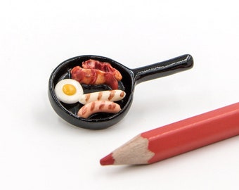 May include: A miniature black frying pan with a white egg, bacon, and sausage inside. The pan is on a white surface next to a red pencil.