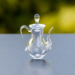 Puede incluir: Una tetera de vidrio transparente con tapa y pico. La tetera está sobre una superficie gris.