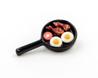 May include: A miniature black frying pan with two fried eggs, bacon, and a tomato slice.