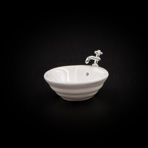 May include: A small, white ceramic bathtub with a silver faucet. The miniature tub has a rounded shape with a ribbed exterior. The faucet is detailed with a cross-shaped handle. The background is solid black.
