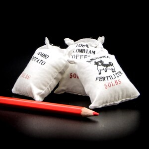 Könnte beinhalten: Drei weiße Stoffsäcke mit schwarzem Aufdruck. Die Säcke sind mit "Idaho Potato 50 LBS", "100% Colombian Coffee 50 LBS" und "Natural Fertilizer 50 LBS" beschriftet.