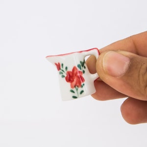 Op de afbeelding: Een kleine witte keramische kan met een rood en groen bloemenmotief. De kan heeft een handvat en een tuit.