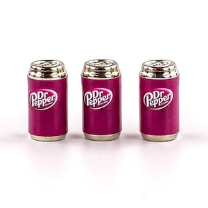 Puede incluir: Tres latas en miniatura de refresco Dr Pepper. Las latas son rosas con tapas plateadas y el logotipo de Dr Pepper en blanco.