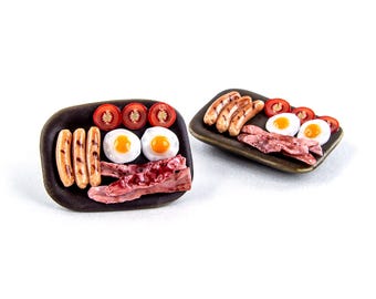 May include: Miniature breakfast plates with fried eggs, bacon, sausages, and sliced tomatoes. The food is arranged on dark brown rectangular plates. The eggs have yellow yolks, and the bacon is reddish-pink.