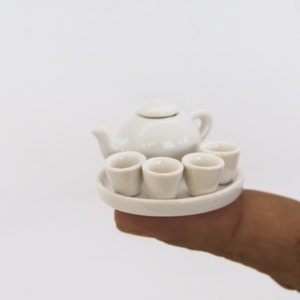 May include: A miniature white ceramic tea set, including a teapot with a lid, four small cups, and a round serving tray. The set is held in a person's hand against a white background. The tea set is a delicate and charming item.