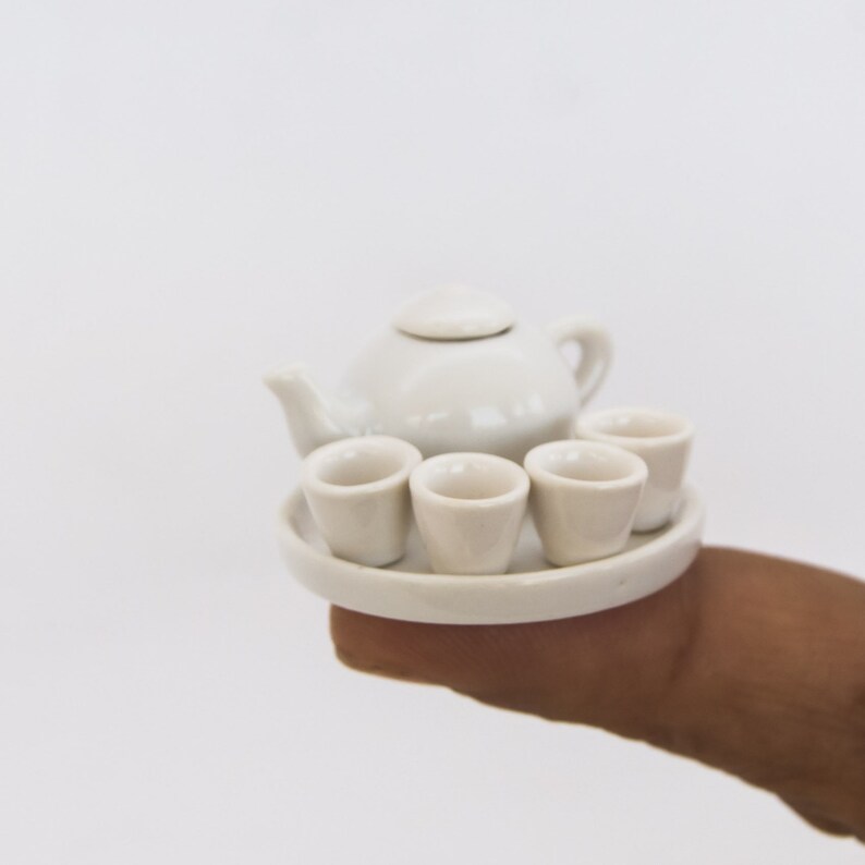 May include: A miniature white ceramic tea set, including a teapot with a lid, four small cups, and a round serving tray. The set is held in a person's hand against a white background. The tea set is a delicate and charming item.