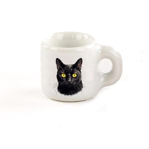 Peut inclure: Une tasse en céramique blanche avec un chat noir aux yeux jaunes imprimé sur le devant.